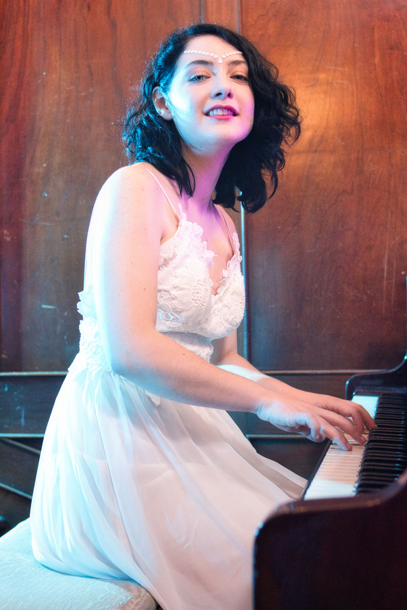 Victoria Daninisa tocando piano de cola con vestido blanco de encaje, mirada a cámara, sesión editorial en interior vintage, Ciudad de México, fotografía por Josué Palma